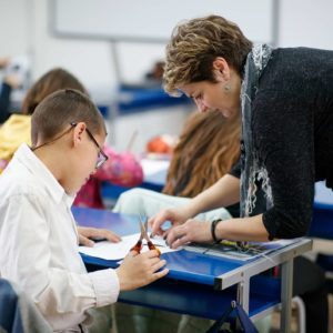 Female teacher working with a special need elementary age boy, she is directing him how to cut paper following pattern